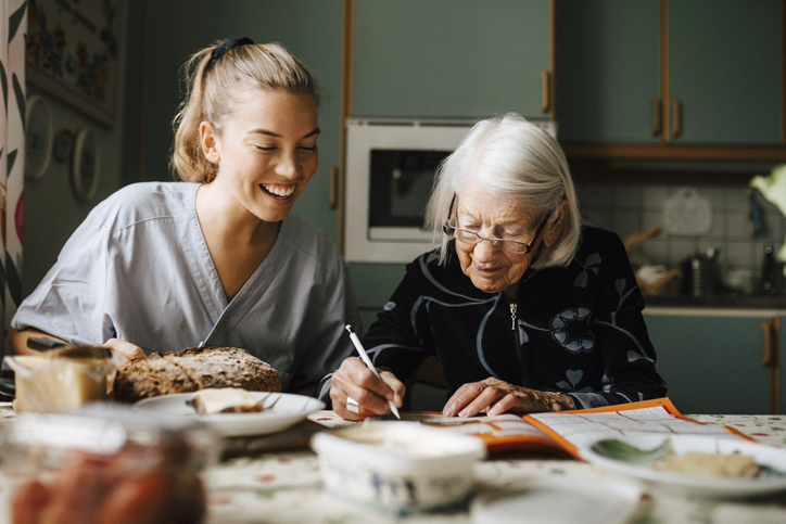 Photo shows Senior woman solving crossword puzzle in book sitting by smiling caregiver in kitchen